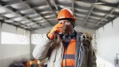 戴着安全帽、穿着工作服的人正站在机库中间，在摄像机前享受一杯咖啡